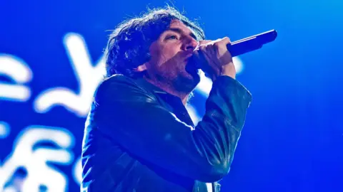Snow Patrol singer Gary Lightbody sings into a microphone on stage wearing a blue jacket. Behind him is a blue screen with white letters.