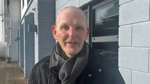 A man looks straight to camera. He is wearing a dark scarf and a black jacket. Behind him there is an office building.