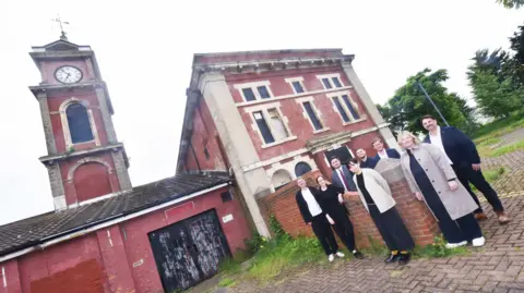Eight members of the engineering and design team standing outside the Old Town Hall. The building is largely painted red and has a clock tower. A number of windows are boarded up. 