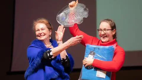 Erin Medcalf Erin Medcalf holding up a glass fish plate and dry robe packaged in her hands next to a woman in a blue fur-trimmed poncho. Erin wears a bright red turtle-neck jumper. They are both smiling.