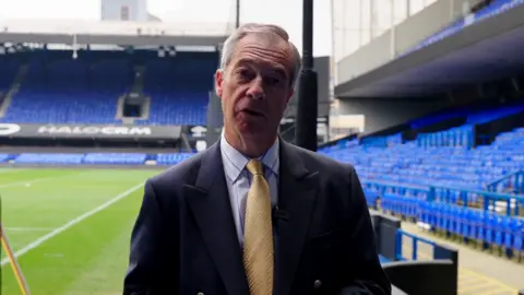 Reform UK Nigel Farage wearing a dark navy suit jacket over a light coloured smart shirt with a yellow tie. He is standing next to the pitch at Portman Road stadium in Ipswich. He is looking directly into the camera.