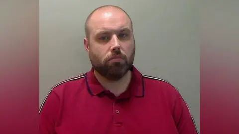 A bald man with a brown beard looks into the camera. He is wearing a red polo shirt.