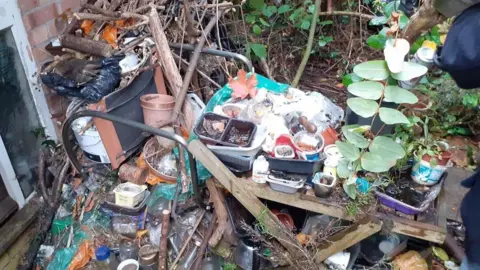 Rubbish at the door of a property. There is garden waste, plant pots, bottles and black bags. 
