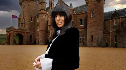 TV Presenter Claudia Winkleman. She has black hair with a long block fringe. She is wearing dark eye makeup and light coloured lipstick. She has a black jacket on with an ornate white shirt underneath. She is standing in front of Ardross Castle.