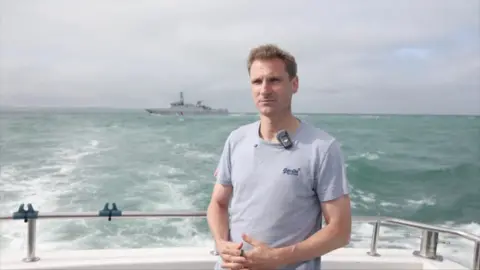 Chris Philp stood on a boat with the French navy patrol boat behind him