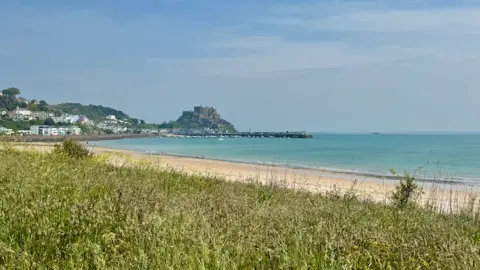 Scenic coastal landscape featuring a sandy beach and grassy dunes in the foreground, calm turquoise waters, and a small town with white buildings along the coastline. Gorey Castle sits atop a hill overlooking the sea under a mostly clear sky with light clouds.