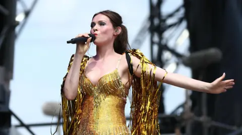 EPA Sophie Ellis-Bextor wearing a gold outfit on stage, covered in gold streamers. she is singing into a microphone and has an outstretched arm.