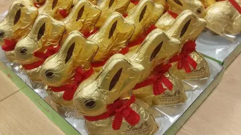A tray has a lot of chocolate bunnies sitting on it. The bunnies are covered in gold foil and have a red ribbon and bells around their necks. Printing on the side reads: "Lindt Gold Bunny"