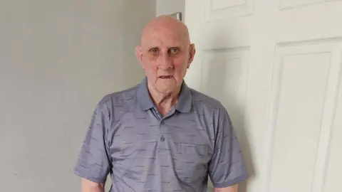 Head-and-shoulders image of Kenneth Forrest. He is bald and wearing a grey polo shirt. He is standing in front of a white door.