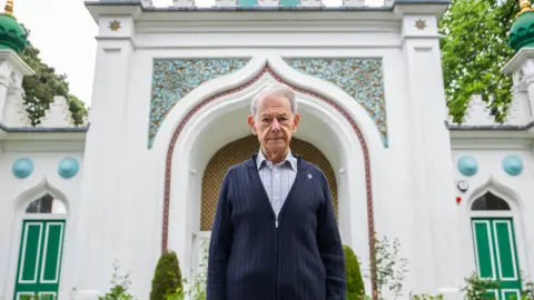 An elderly man wearing a grey shirt and dark blue cardigan. He is facing the camera and stood outside a white mosque