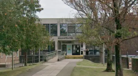 A single-storey building set back from the street, with a sign above the entrance reading “Magistrates Court”. A paved walkway with trees on either side leads directly to the front doors, flanked by metal railings on a sloped access ramp.