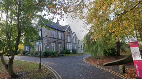 A large stone-built house with a black roof and large windows. There are many trees surrounding the house and a small road leading up to it.