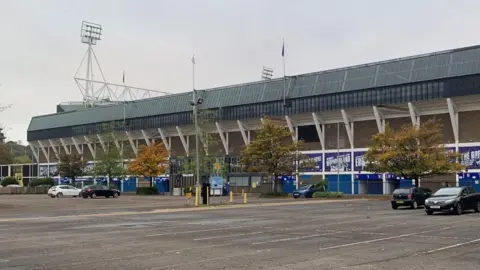 Mark Bulstrode/BBC Portman Road, as seen from New Portman Road car park. The two-tier stadium can be seen in the background, with its black roof and white supporting columns outside. In the foreground are cars parked in a car park on a cloudy day.
