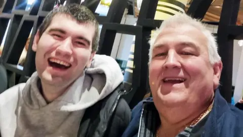 Aaron and his dad Glynn captured side by side with wide, open smiles - as if caught laughing. Aaron, on the left, is wearing a grey hoodie and his dad is wearing a dark blue jacket.
