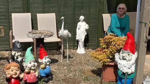 Vikki Henderson Vikki Henderson, wearing sunglasses, is pictured sitting on a chair surrounded by gnomes in a small, gravelled back garden. In the background is a wooden fence, two deckchairs, a bird bath and a white heron statue.