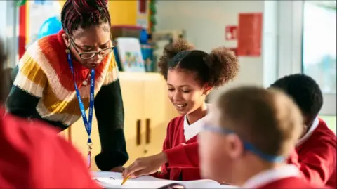 A teacher with red and black braids placed in a bun is leaning over a table talking to pupils. A girl has two afro buns on the side of her head and is wearing a white polo shirt under a red cardigan. The teacher is wearing glasses and a red, orange, yellow, green and white wool jumper.