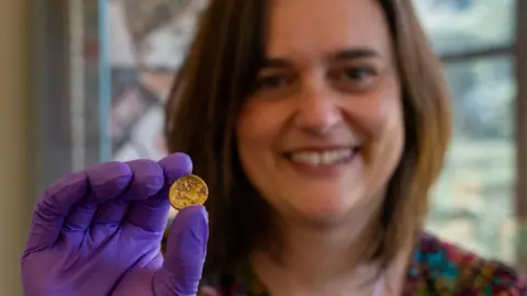Fountain Media An out of focus Claire Willetts, who has shoulder-length light brown hair and is smiling, holding up a gold Iron Age coin which is in focus. She is wearing purple gloves.