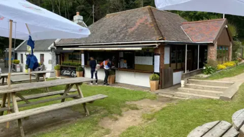 An outdoor cafe with people queuing at the front, outdoor tables with umbrellas on them.