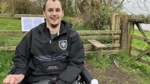 A man in a wheelchair in beside an wooden fence stile along the green trail