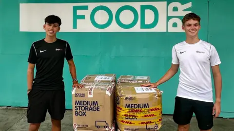 Alfie Watts Two young men are stood next to a waist high pile of cardboad boxes while wearing shorts