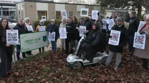 A group of campaigners hold protest placards in the grounds of Great Alne School. They are dressed in coats for what looks like a cold day.