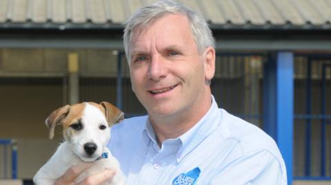 Neil Edwards holding a puppy which has white fur and brown ears as Mr Edwards smiles at the camera. He is wearing a blue shirt with the logo for the Blue Cross charity on it and stands in front of a building with blue posts and a grey roof