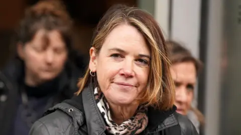 A woman with brown hair with blonde highlights leaves a building with two other women in the background. The woman is wearing a black jacket and a leopard print scarf. 