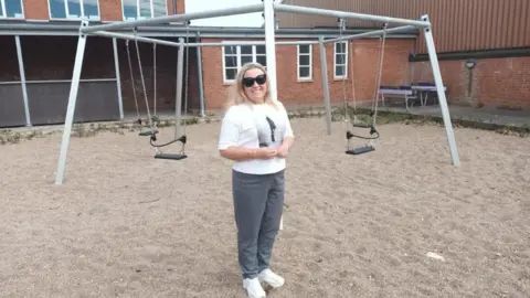 A woman with blonde hair and sunglasses stands in front of a bare playground made of sand and dilapidated swings, in front of an old school building.