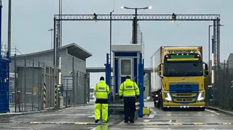 Officials await the arrival of an HGV at a border check area. They are both in yellow hi-vis. A lorry is coming through the port. 