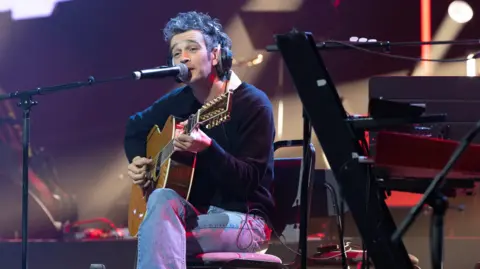 John Millard Photography Matty Healy sitting on a chair playing an acoustic guitar and singing into a microphone. He has dark hair which is sticking up on the top and has large over-the-ear headphones on. He is wearing a dark jumper and light blue jeans and his legs are crossed. He is looking ahead of him which is to the left of the camera.