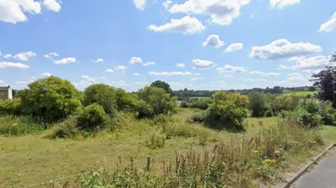 An area of green land with trees and bushes, with a road running through the bottom right corner of the image. The sky is blue with some clouds.