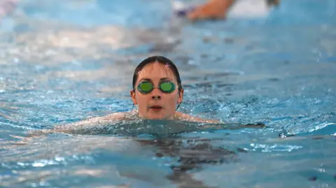 A woman with dark hair in goggles is swimming in an indoor pool