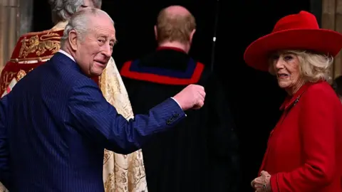 The King and Queen turn around to look behind them. The King holds a hand up while the Queen smiles