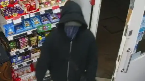 A still image of the offender taken from the store's CCTV. He is wearing a dark coloured hoodie and has a blue piece of material covering his face. 