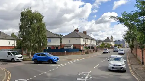 Google maps street view of St Paul's Road and the junction for Folly Lane in Cheltenham. There are cars on the road and houses on the street.