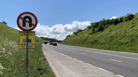 A30 Hayle bypass in Cornwall. Vehicles are travelling along the road. Green banks lined either side of the carriageway. U-turn signage has been installed, including an Operation Snap sign which says "U-turn on dashcam? Submit to Op Snap"