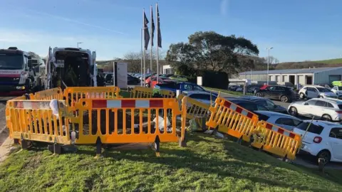 A site on an industrial estate. In the middle is a a yellow temporary barrier on a grass verge. Cars and lorries can be seen to side the side and in the background.