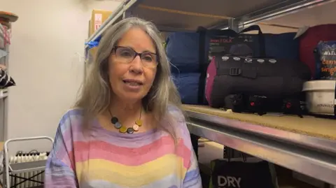 A woman with glasses and a striped top and a chunky necklace. Behind her are shelves with camping accessories.