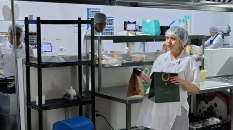 Foodology A Foodology worker in cooking work clothes carries two bags of food. Behind are other workers in the kitchen.