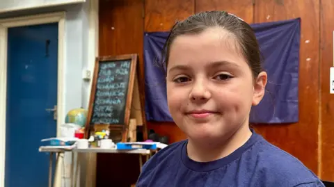 A nine-year-old girl is smiling and looking towards the camera. She has brown hair and a blue t-shirt.