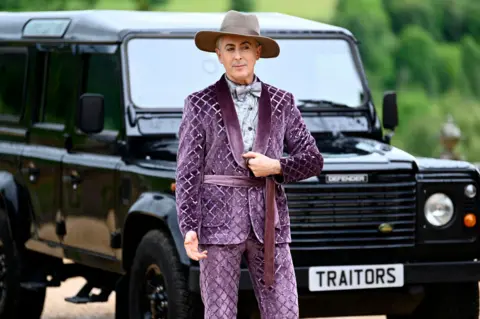 BBC/Peacock TV/Euan Cherry Alan Cummings in the TV programme The Traitors US, standing in front of the Traitors 4x4 in a mauve purple velvet suit with a diamond sequin pattern, contrasting purple velvet collar and tie belt, grey silk shirt and bow tie and a wide-brimmed mink coloured fedora hat.