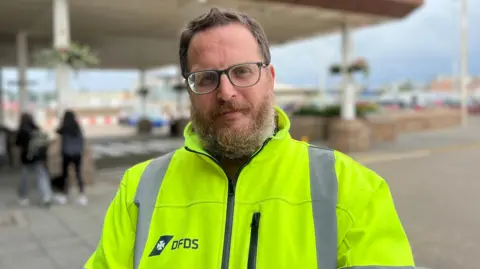 A man called Chris Parker who works for ferry firm DFDS. He is wearing a yellow high-vis coat with the company's logo on it. He has trimmed brown hair and a greying beard which covers his lower face. Parker is wearing glasses.