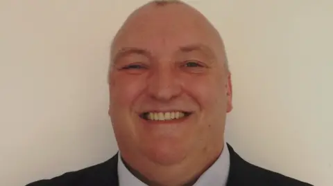 St Gregory's Catholic Club Headshot of Mark. He is smiling at the camera, in front of a pale background. He has short shaved head. 