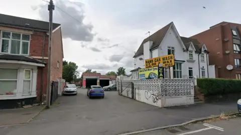 A flat tarmacked area with at least three cars on it is photographed. A yellow sign next to the area reads: "Hand car wash and wax £5."