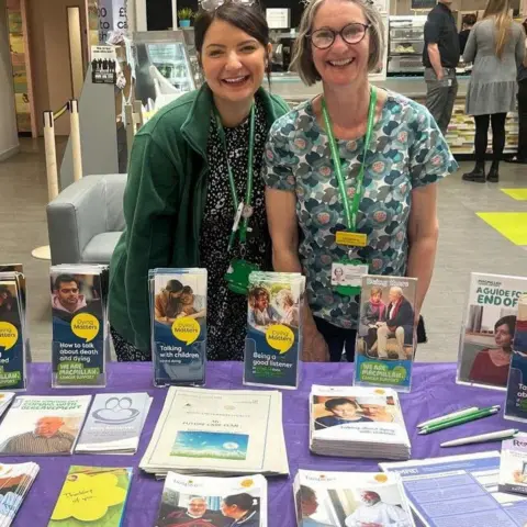 NHS Two women smiling. One has dark hair and wears green fleece. The other has light hair and wears a patterned blue dress. They both have lanyards around their necks and are standing behind a table with leaflets on it.