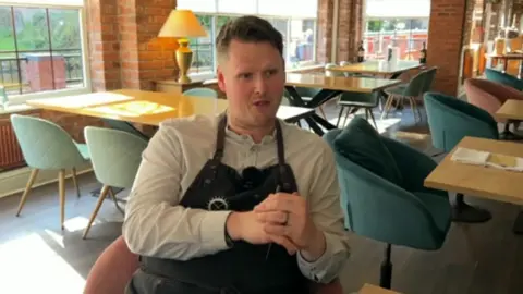 A man in a long sleeve top and dark apron, sitting on a chair in a restaurant, his hands clasped together