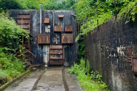 BBC The exterior of a cement building, there are rusty boxes and doors on the front. there is a rail track going all the way down the lane and into the building. There is a storm drain on the left of the tracks and there is overgrowth on all sides.