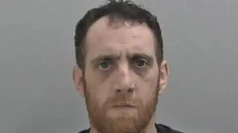 West Mercia Police Bearded man with a cut on his nose looks directly into the camera against a grey wall.