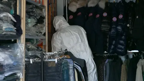 Man in white suit examines imitation Canada Goose fur-hooded parka coats.