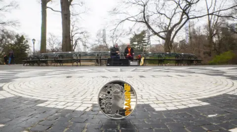 The Royal Mint A silver coin with a portrait of John Lennon on it, with lettering that reads John Lennon on one side, and the words 'Imagine' in yellow, on the opposite side. The coin is sat upright on cobblestones in a park, with two men sitting on a bench with a guitar in the background